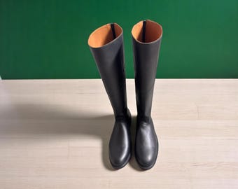 以下が含まれることがあります： 滑らかな黒色の乗馬ブーツのペア。内側は明るい茶色です。ブーツは緑と明るい木目の背景に立っています。ブーツはクラシックな乗馬スタイルです。
