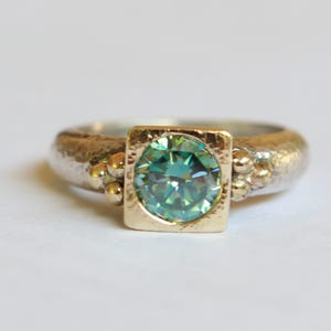 Peut inclure: Une bague en argent avec un anneau texturé et un sertissage carré en or. Le sertissage contient une pierre précieuse ronde de couleur teal. De petites perles d'or ornent les côtés du sertissage de la pierre.