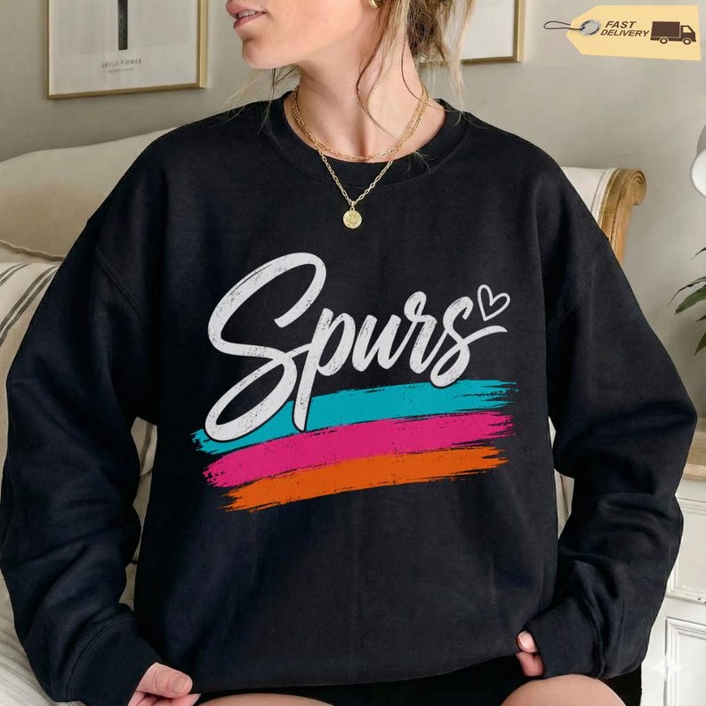 May include: Black sweatshirt with the word "Spurs" in white script, above a horizontal brushstroke design in teal, pink, and orange. A small heart is next to the word "Spurs". The sweatshirt is a casual apparel item.
