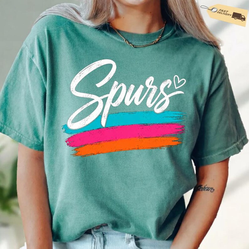 May include: Teal-colored t-shirt with the word "Spurs" in white script, above three horizontal brushstrokes in blue, pink, and orange. A small heart is next to the word "Spurs". The shirt has short sleeves.