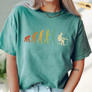 K&ouml;nnte beinhalten: Ein kurz&auml;rmeliges T-Shirt in Blaugr&uuml;n mit einem Grafikdruck der menschlichen Evolution in Orange-, Gelb- und Gr&uuml;nt&ouml;nen. Das Design zeigt die Entwicklung vom Primaten bis zum Menschen, der an einem Schreibtisch an einem Laptop arbeitet.