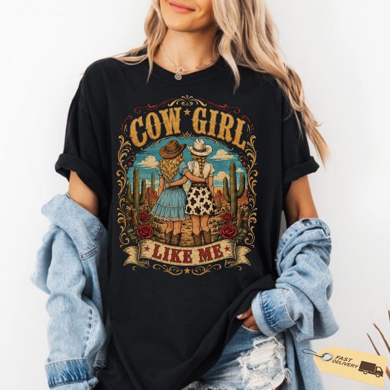 May include: A black short-sleeve t-shirt featuring a graphic print of two figures in cowboy hats standing in a desert with cacti. The text COW GIRL LIKE ME is printed in gold lettering. The design includes ornate scrollwork and red roses.