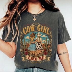 May include: A charcoal grey t-shirt featuring a graphic print of two figures in cowboy hats and dresses standing in a desert landscape with cacti and red roses. The text COW GIRL LIKE ME is printed in gold and red lettering around the illustration.
