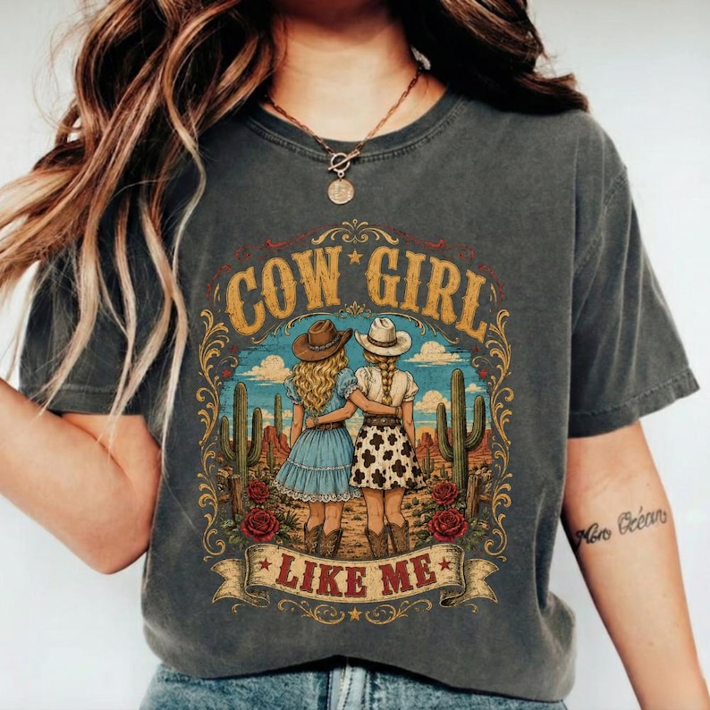 May include: A charcoal grey t-shirt featuring a graphic print of two figures in cowboy hats and dresses standing in a desert landscape with cacti and red roses. The text COW GIRL LIKE ME is printed in gold and red lettering around the illustration.