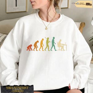 K&ouml;nnte beinhalten: Ein wei&szlig;es Langarm-Sweatshirt mit einem mehrfarbigen Grafikdruck, der die Evolution der Menschheit darstellt, beginnend bei einem Primaten und fortschreitend &uuml;ber verschiedene Stadien bis hin zu einer Person, die an einem Schreibtisch am Laptop arbeitet.