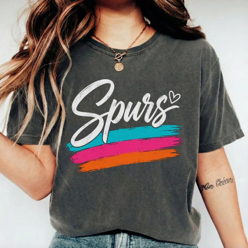 May include: Dark gray t-shirt with the word "Spurs" in white script, a heart, and horizontal stripes in turquoise, pink, and orange. The shirt is a casual style.