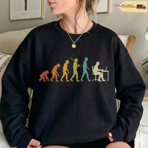 K&ouml;nnte beinhalten: Ein schwarzes Langarm-Sweatshirt mit einem mehrfarbigen Grafikdruck, der die Evolution der Menschheit darstellt, beginnend bei einem Primaten und fortschreitend &uuml;ber verschiedene Stadien bis hin zu einer Person, die an einem Schreibtisch am Laptop arbeitet.