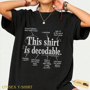 Op de afbeelding: Een zwart t-shirt met korte mouwen en de witte tekst This shirt is decodable. Het ontwerp bevat diverse taalkundige annotaties met pijlen die naar specifieke woorden wijzen, waarbij concepten zoals gesloten lettergrepen en digrafen worden uitgelegd.