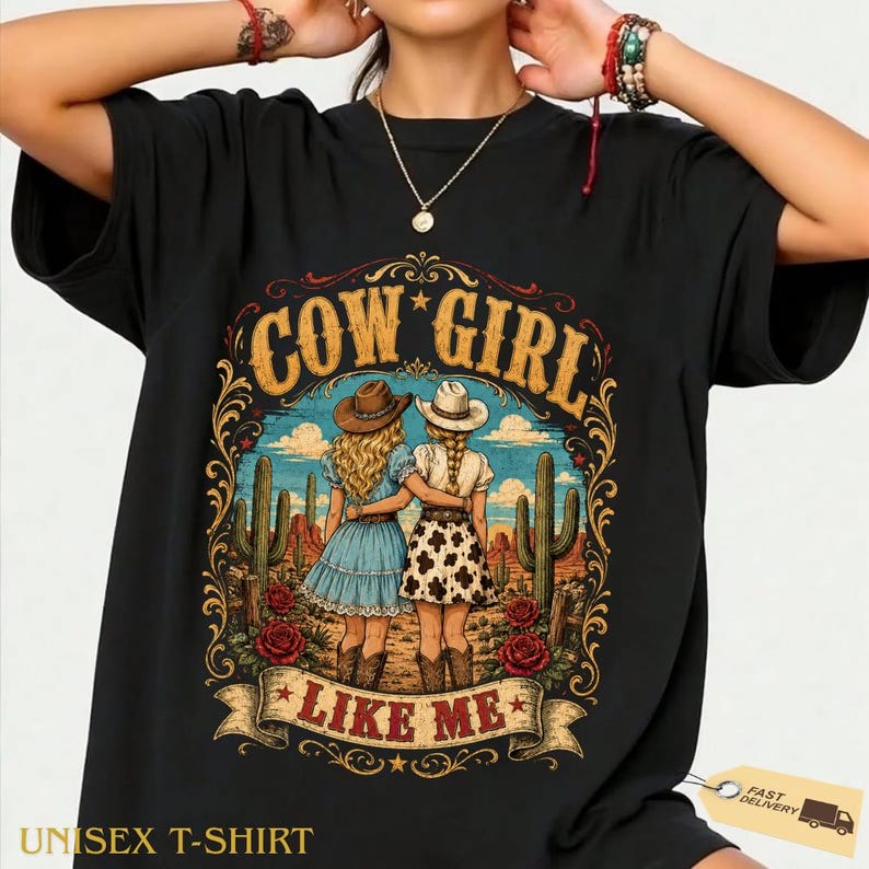 May include: A black short-sleeved t-shirt featuring a graphic print of two individuals in cowboy hats standing in a desert landscape with cacti and red roses. The text COW GIRL LIKE ME is printed in a decorative font above and below the illustration.