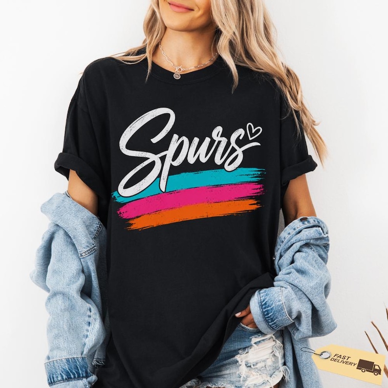 May include: Black t-shirt with the word "Spurs" in white script, a teal, pink, and orange brushstroke design, and a small heart. The shirt is a casual style.