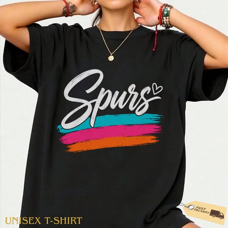 May include: Black unisex t-shirt with the word "Spurs" in white script above horizontal stripes of turquoise, pink, and orange. A small heart is next to the word "Spurs". The text "UNISEX T-SHIRT" is at the bottom.