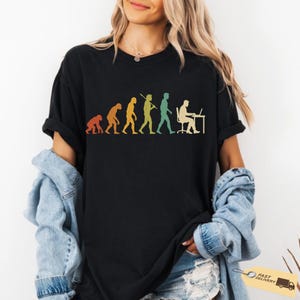 May include: A black short-sleeve t-shirt featuring a colourful graphic print of human evolution, starting from a primate and progressing to a person sitting at a desk with a computer. The design uses a retro-inspired colour palette of orange, yellow, and green.
