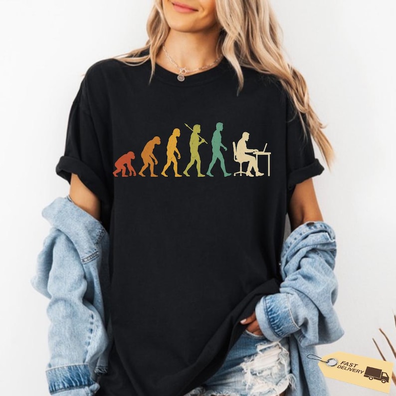 K&ouml;nnte beinhalten: Ein schwarzes Kurzarm-T-Shirt mit einem farbenfrohen Grafikdruck der menschlichen Evolution, der von einem Primaten bis hin zu einer Person reicht, die an einem Schreibtisch mit einem Computer sitzt. Das Design verwendet eine Retro-Farbpalette.
