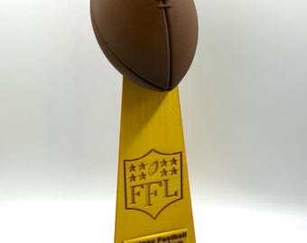 May include: A brown football trophy with a yellow base and stand. The trophy features the text "FFL" and "Fantasy Football Champion". The football is positioned at the top of the trophy.