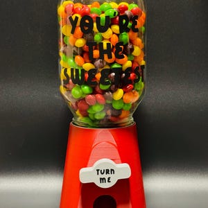 May include: A red gumball machine with a clear glass jar filled with colorful candies. The jar has the text "YOU'RE THE SWEETEST" in black. The machine has a white "TURN ME" sign.