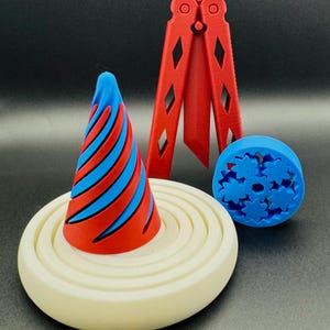 May include: A set of three 3D printed objects: a red and blue cone, a red butterfly knife, and a blue gear. The cone is sitting on a white circular base with concentric rings.