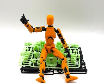 May include: An orange articulated figure with black joints and hands, posed with one arm raised. The figure is sitting in front of a pile of green plastic model kit parts. The figure has the number 13 on its back.
