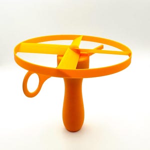May include: An orange and yellow toy helicopter with a handle and a circular ring. The toy has a propeller design with four blades and a small loop on the handle. The toy is made of plastic and is designed for children.