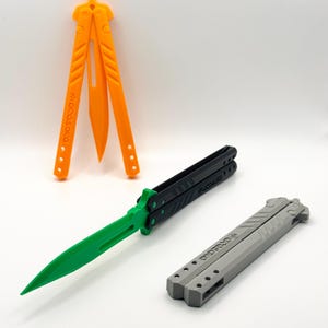 May include: Three butterfly knives, one orange, one black and green, and one grey. The knives are closed and laid out on a white surface.