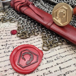 Könnte beinhalten: Ein rot lederumwickelter Schwertknauf und ein Messing-Wachssiegel mit einem Skorpion-Design. Das Siegel befindet sich neben einem roten Wachssiegel mit demselben Skorpion-Design. Kettenhemden und ein Rubin sind ebenfalls sichtbar.