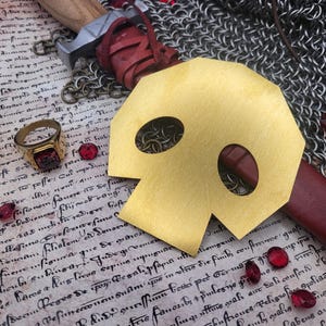 May include: A gold skull mask with eye and nose cutouts lies on a parchment-like surface. A red-stoned ring, red gems, a sword, and chainmail are also present. The scene suggests a medieval or fantasy theme.