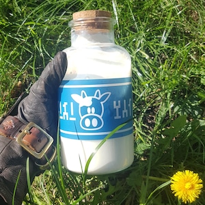 Puede incluir: Una botella de vidrio transparente llena de un líquido blanco, sellada con un tapón de corcho. La botella tiene una etiqueta azul con una vaca de dibujos animados y texto en idioma extranjero. La botella se sostiene con un guante de cuero negro, sobre hierba verde y un diente de león amarillo.