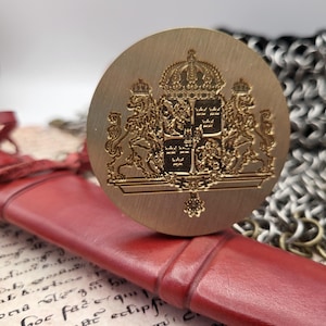 Puede incluir: Un sello redondo de color dorado con un escudo de armas en relieve detallado, que incluye una corona y leones heráldicos. El sello descansa sobre una superficie de cuero rojo, con una gema de color rubí y cota de malla en el fondo.