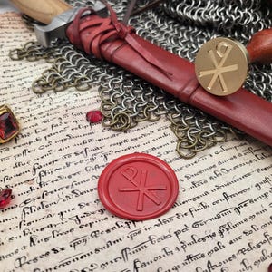 Könnte beinhalten: Eine Ansammlung historischer Artefakte, darunter ein goldener Ring mit rotem Edelstein, eine rote Lederscheide, ein Messingstempel und ein rotes Wachssiegel. Die Gegenstände sind auf einer pergamentartigen Oberfläche mit handschriftlichem Text und Kettenhemden angeordnet.