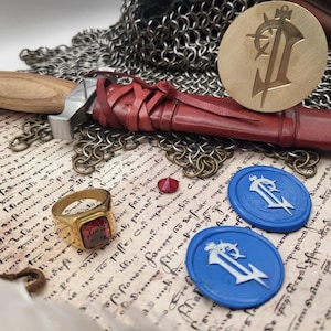 May include: An assortment of medieval-themed items, including a sword with a wooden handle and red leather sheath, a gold ring with a red stone, and two blue wax seals. A brass seal and chainmail are also visible.