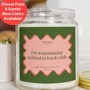 Pode incluir: Uma vela de vidro com um rótulo verde e uma caixa de texto rosa com bordas onduladas que diz "I'm waaaaaaaay behind in book club". A vela tem 255g e é feita de cera natural vertida à mão. Um círculo vermelho diz "Escolha entre 8 fragrâncias! Mais cores disponíveis!"