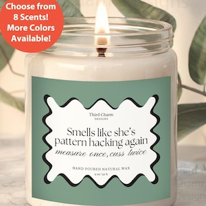 May include: A green and white candle with the text "Smells like she's pattern hacking again measure once, cuss twice" and "Third Charm Designs" on the label. The candle is hand-poured natural wax and is 9 oz/50 h.