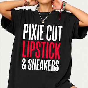 Puede incluir: Camiseta negra con las palabras "PIXIE CUT LIPSTICK & SNEAKERS" en letras blancas y rojas. La camiseta tiene mangas cortas y cuello redondo.