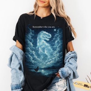 Puede incluir: Camiseta negra con una ilustración surrealista de un dinosaurio formado por nubes, una luna llena y una gallina. El texto "Remember who you are" está impreso encima de la imagen. La camiseta es de un material suave.