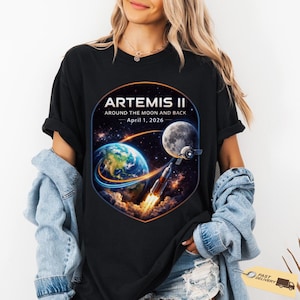 Puede incluir: Camiseta negra con un diseño espacial colorido. El diseño incluye la Tierra, la Luna, un cohete y una nave espacial, con el texto "ARTEMIS II AROUND THE MOON AND BACK - April 1, 2026".