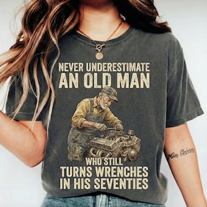 May include: Dark gray t-shirt with the text "NEVER UNDERESTIMATE AN OLD MAN WHO STILL TURNS WRENCHES IN HIS SEVENTIES." The graphic shows an older man working on an engine. The shirt is a vintage style.