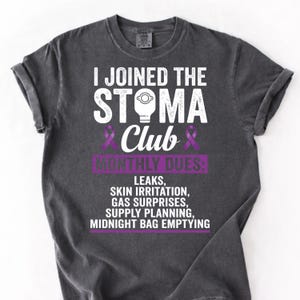 May include: Dark gray t-shirt with the text "I JOINED THE STOMA Club" in white and purple. The shirt lists "MONTHLY DUES" including "LEAKS, SKIN IRRITATION, GAS SURPRISES, SUPPLY PLANNING, MIDNIGHT BAG EMPTYING".