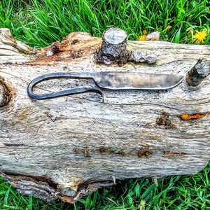 Könnte beinhalten: Ein handgeschmiedetes Messer mit gebogenem Griff liegt auf einem verwitterten Baumstamm. Die Klinge ist silberfarben und der Griff ist schwarz. Der Baumstamm ist hellbraun mit dunkelbraunen Knoten. Der Hintergrund ist grünes Gras und gelbe Löwenzahne.