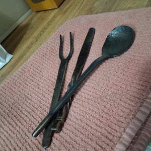 May include: Hand-forged metal cutlery set including a fork, knife, and spoon. The utensils have a dark, rustic finish and are arranged on a pink towel. The spoon has a rounded bowl, and the knife has a twisted handle.