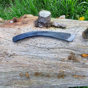 Puede incluir: Una herramienta curva de color gris oscuro con un borde afilado, posiblemente un cuchillo de tracción o un instrumento de carpintería similar, descansa sobre un tronco desgastado. El tronco está rodeado de hierba verde y algunas flores amarillas.