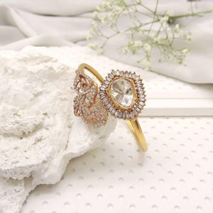 May include: A gold-toned cuff bracelet featuring a large, central pear-shaped stone surrounded by a halo of small, clear baguette-cut stones. One side of the bracelet displays an intricate, openwork floral design encrusted with tiny clear stones.