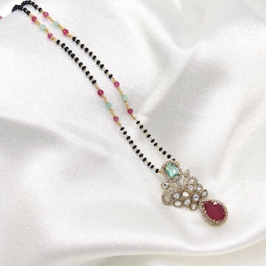 May include: A gold chain necklace featuring small black beads interspersed with pink and light green beads. The pendant is an ornate gold design set with a rectangular light green stone, small clear crystals, and a large teardrop-shaped red stone.