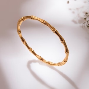 May include: A gold-colored bangle bracelet with a bamboo design. The bracelet is made of a shiny, metallic material and has a smooth, polished finish. The bamboo-style design is visible throughout the bracelet.