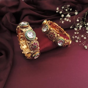 May include: Two gold-colored bangles with intricate floral and geometric designs. Each bangle features large, clear, round stones, surrounded by green and red accents. The bangles are displayed on a deep red satin surface, with white flowers in the background.