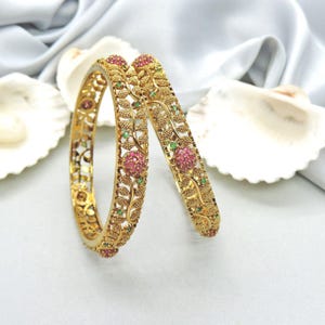 May include: Two gold bangles with intricate floral and leaf designs. The bangles are adorned with pink and green gemstones, creating a vibrant contrast against the gold. The jewelry is displayed on a white surface with seashell accents.