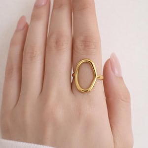 May include: A gold-colored ring with an irregular oval design. The ring is worn on a finger, and the hand is resting on a white surface. The ring's design is a continuous loop, creating an open space in the center.