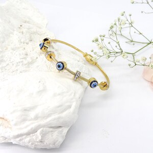 May include: Gold-toned bangle bracelet featuring blue evil eye charms and clear crystal accents. The bracelet has a flexible, woven design and is displayed on a white, textured surface. Delicate white flowers and a pink petal are in the background.
