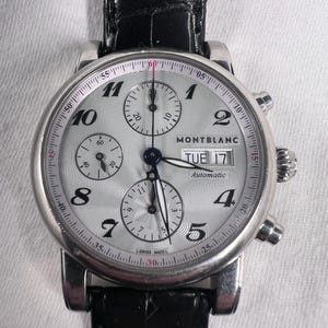 May include: A silver Montblanc automatic wristwatch with a black leather band. The watch face is silver with black numerals and three sub-dials. The date reads Tuesday 17. The watch has two side buttons and a crown.