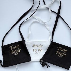 Könnte beinhalten: Drei Gesichtsmasken-Taschen: zwei schwarze und eine weiße. Die schwarzen Taschen sind mit "Team Bride 2026" in Gold bestickt. Auf der weißen Tasche steht "Bride to be 2026". Jede Tasche hat einen verstellbaren schwarzen oder weißen Riemen.