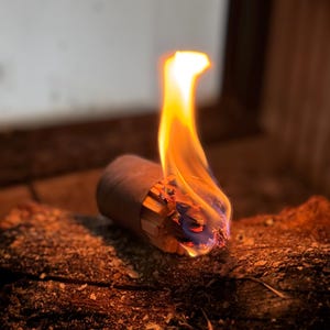 Könnte beinhalten: Nahaufnahme eines brennenden Objekts mit hellen Flammen. Die Flammen sind orange und gelb, mit einem Hauch von Blau an der Basis. Das Objekt scheint ein aufgerolltes Stück Papier oder Pappe zu sein, das auf einer dunklen, strukturierten Oberfläche liegt.