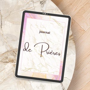 Peut inclure: Une tablette numérique affiche une couverture de journal avec les mots "Journal de Prières". La couverture présente un motif marbré avec des accents dorés, une croix et une bordure rose. La tablette est posée sur une table en marbre.